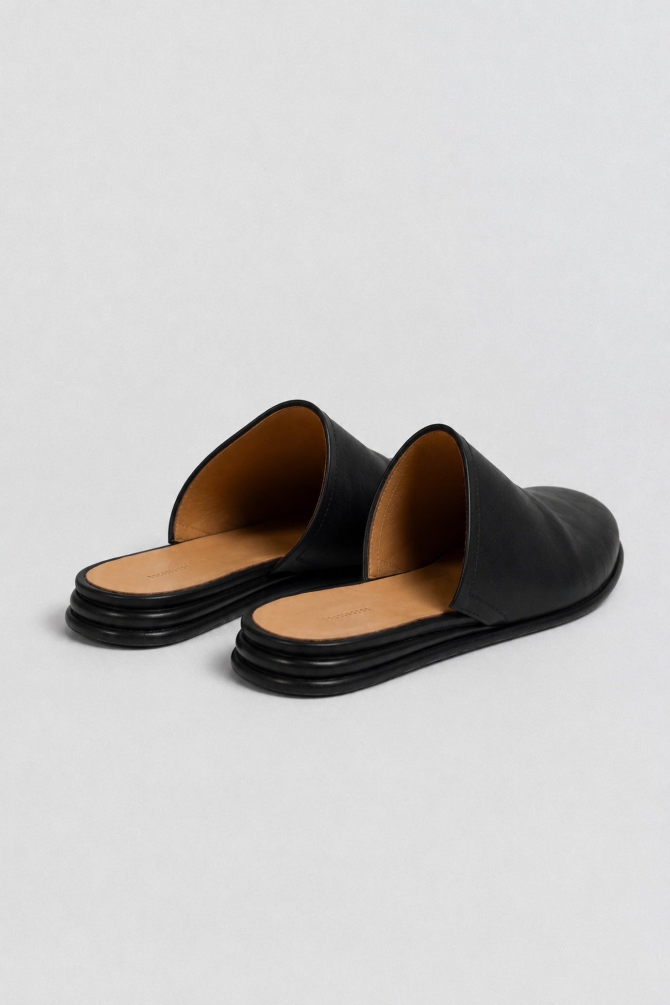 A pair of black leather mule slide sandals with a tan interior lining and black multi-layered soles, shown on a light gray background.