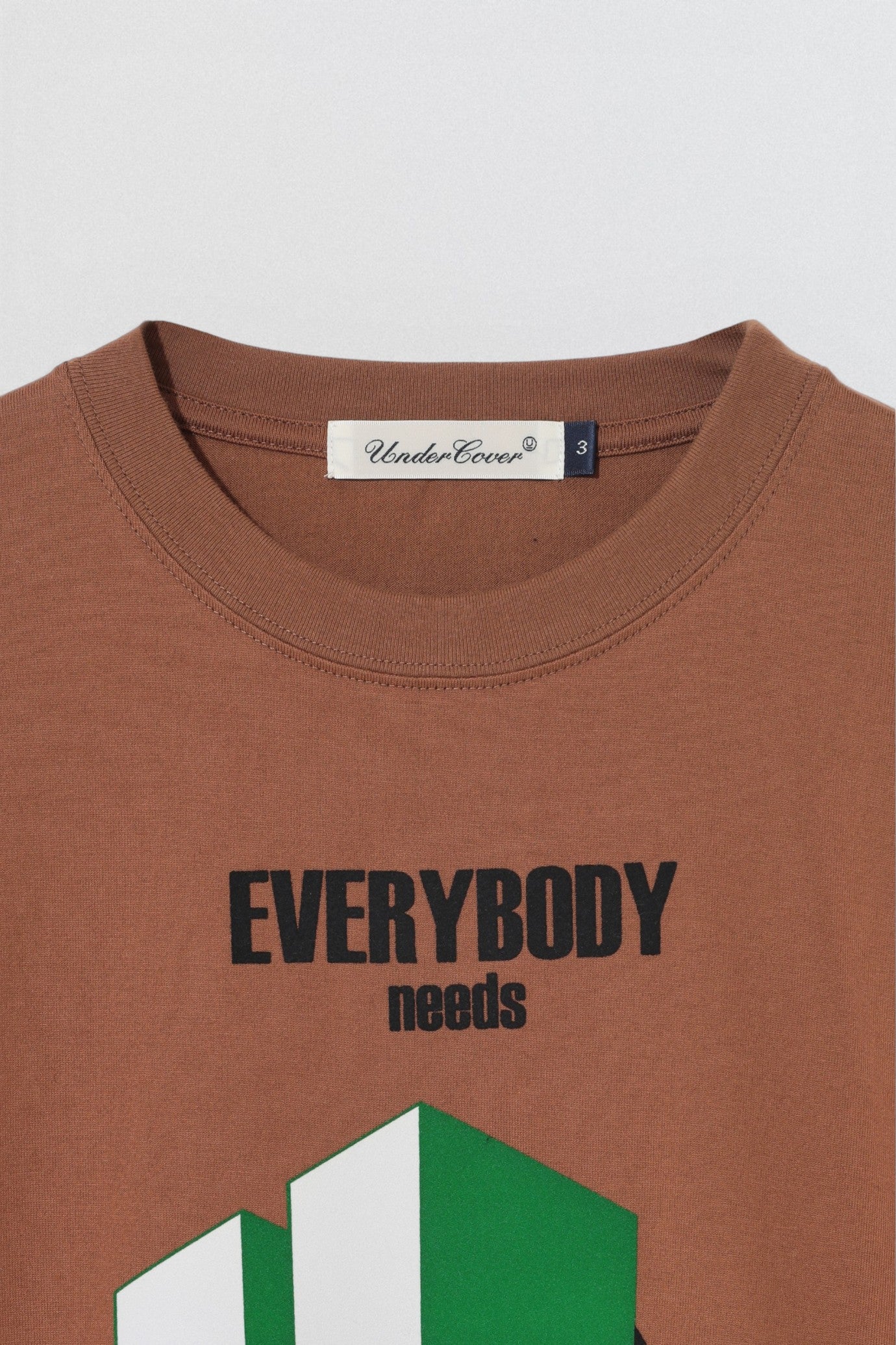 Undercover SS26 Brown t-shirt with 'EVERYBODY needs' text and green geometric design, brand label visible.