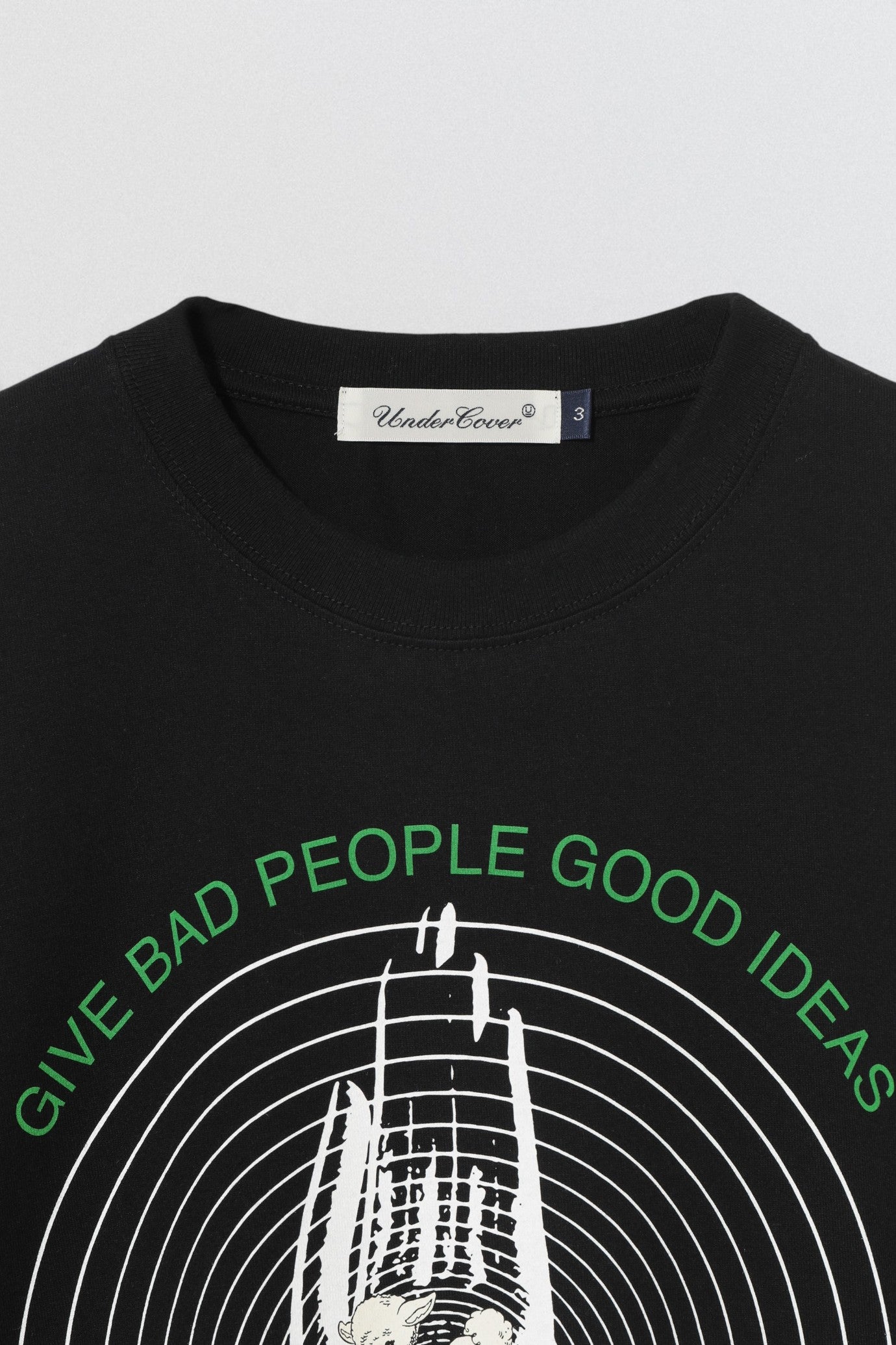 Undercover SS26 Give Bad People Good Ideas Black t-shirt with green text and graphic design on a light gray background
