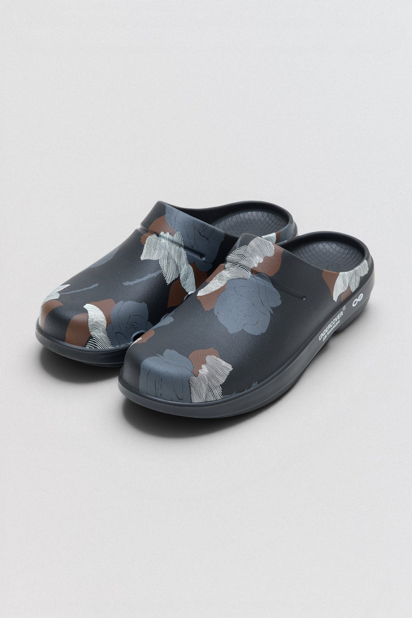 Undercover x OOFOS Pair of gray clogs with floral pattern on a light gray background