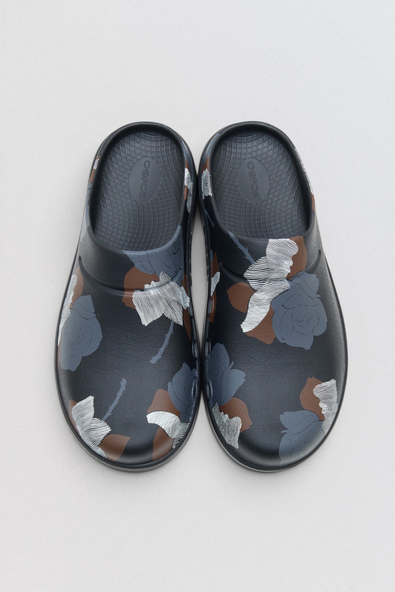 Undercover x OOFOS Pair of black clogs with floral pattern on a light gray background